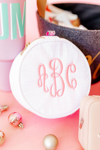 A white round cosmetic bag with a monogram in pink letters, displayed on a pink surface surrounded by various items including a black brush, ornaments, and a gold object.
