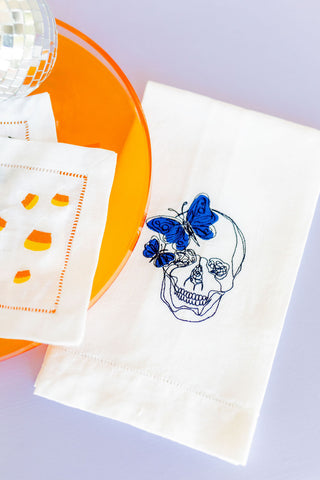 A white embroidered guest towel with a navy blue butterfly and a skeleton design, displayed on an orange plate.