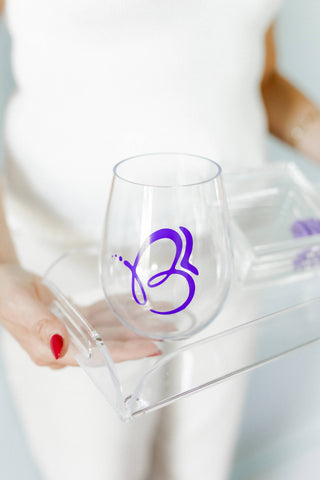 Clear wine glass with a purple logo held by a person with red nail polish. | Whatsoever Things | Dallas, TX | Personalized Apparel, Decor and Gifts for Men and Women | Embroidery and Monogramming 